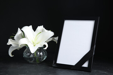 Funeral photo frame with ribbon and white lilies on black table against dark background. Space for...