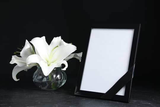 Funeral photo frame with ribbon and white lilies on black table against dark background. Space for design