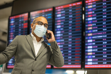 Young businessman talking on phone in the airport. Wearing mask. traveling during pandemic must be social distancing. He standing at information flight timetable waiting plane at airport terminal.