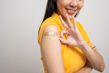 Vaccination. Young beautiful asian woman in yellow shirt getting a vaccine protection the coronavirus. Smiling happy female showing arm with bandage after receiving vaccination. Showing ok sign. - obrazy, fototapety, plakaty