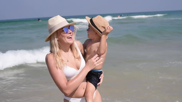 Mom in white bikini and sunglasses holds son on hands. Woman and boy in hats on seashore. Dancing with happy smiling. Sunny summer, vacation on sea,  travel, outdoors.