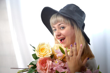 Happy beautiful woman with long blond hair and in black hat posing with flowers in studio on white background