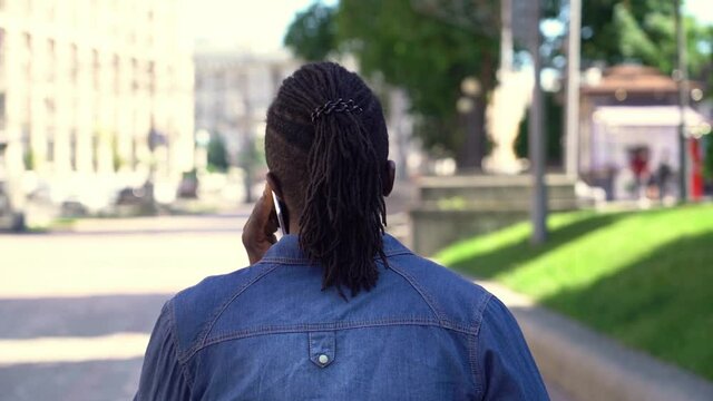 Back view of African hipster man with stylish hairstyle walking on the street talking on mobile phone, slow motion 
