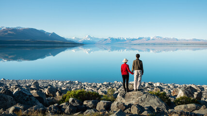 Couple standing on the shore of Lake Pukaki, holding hands and watching Mt Cook reflected in the clear waters, South Island, New Zealand