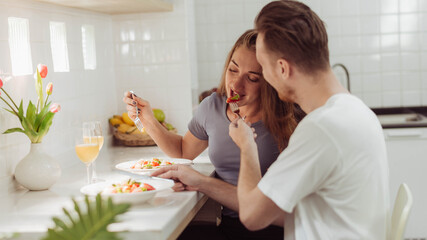 Young man giving strawberry to his girlfriend while cooking together in the kitchen. Happy couple love cooking together at home.