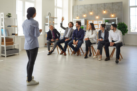 Young Man In Audience Raises Hand To Ask Speaker A Question. Small Group Of People Sitting In Row In Modern Office Space Listening To Business Trainer, Corporate Psychologist Or Personal Growth Coach