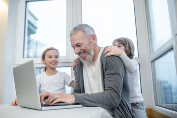 Gray-haired bearded man working on laptop while his kids interfering him - obrazy, fototapety, plakaty