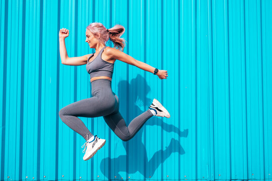 Fitness smiling woman in grey sports clothing with pink hair. Young  beautiful model with perfect body.Female posing in the street near blue  wall.Cheerful and happy. Jumping and running Stock Photo | Adobe