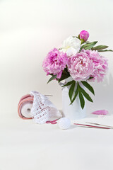 Summer needlework and crochet. Bouquet of fresh pink peonies and travel bag with balls of cotton yarn for crocheting lace on white background. DIY concept. High key, soft focus. Close-up, copy space