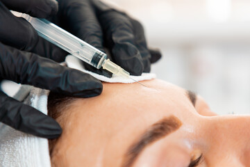 Close-up of female's head getting injection at her forehead in the cosmetology salon. Cosmetologist in latex gloves with syringe injects a medicine. Concept of plastic surgery and face lifting