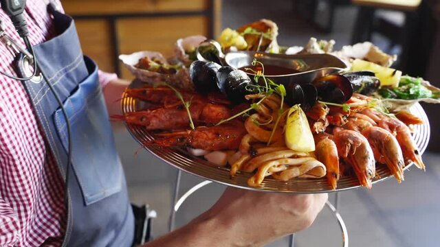 Waiters carries mixed seafood set, lobster shrimp, king prawns, mussels, oysters and squids served on large salver plate for luxury restaurant visitors. Garcon brings traditional meditarian dish.