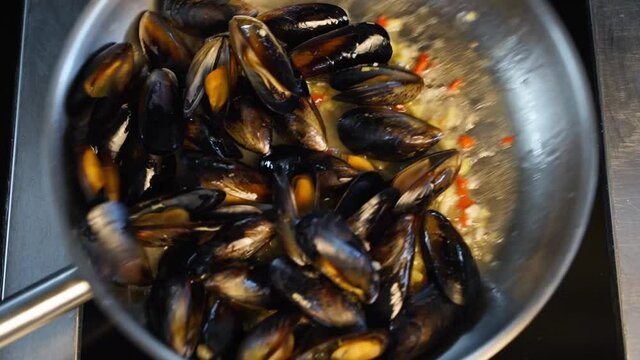 Cooking delicious fresh mussels on hot pan in luxury restaurant. Chef shakes and mixes mussels and greens on a grill. Traditional mediterranean cuisine. Slow motion scene.