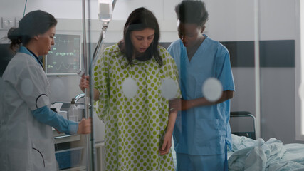 Black asisstant with practitioner doctor helping patient to stand up from bed holding intravenous IV fluid drip bag after medical surgery in hospital ward. Specialist checking cure expertise