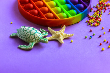 Trendy silicone anti-stress toy Pop it in rainbow colors on a violet background flatly. Toy turtle, starfish, foam beads scattered on a table. Summer holiday concept. Space for text. Fine motor skills