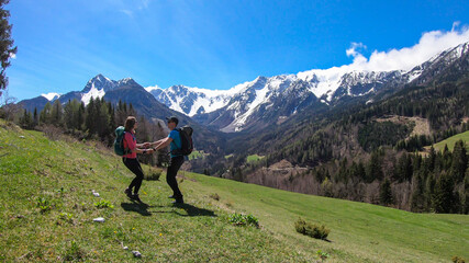 A couple with a big hiking backpacks dancing with the panoramic view on Baeren Valley in Austrian Alps. The highest peaks are snow-capped. Lush green pasture. Clear and sunny day. High mountain chains