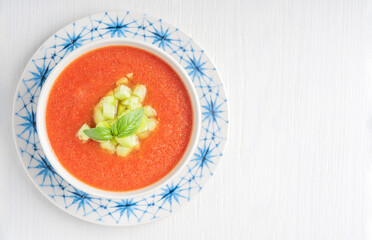 Top view of spanish gazpacho cold refreshing tomato soup made of raw blended vegetables served in bowl on plate with basil leaves and cucumber cubes on white wooden background. Image with copy space