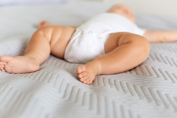Newborn baby legs, faceless infant wearing white bodysuit lying on the gray blanket in bed,...