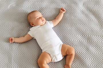 Charming cute baby lying on a bed, sleeping in a bright room on grey blanket, laying with spreading hands, afternoon sleep indoor.