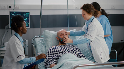 Medical team monitoring heartbeat pulse of sick man during respiratory emergency working in hospital ward. Patient sitting in bed while doctor putting oxygen mask analyzing breath condition