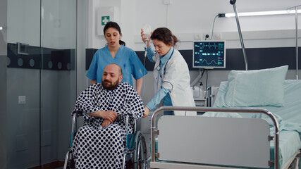 Fototapeta premium Medical team helping sick patient with leg fracture putting in wheelchair going on physiotherapy recovering in hospital ward. Doctor consulting patient checking respiratory expertise