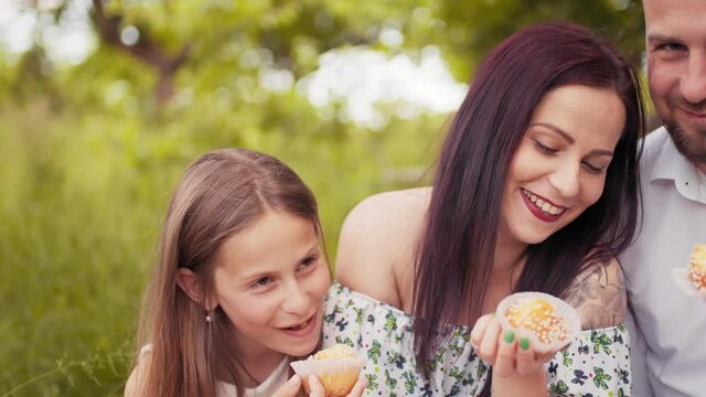 Smiling parents with two cute daughters in summer clothes eating homemade cupcakes during picnic time at green garden. Concept of people, leisure and relaxation. Parents with kids eating cupcakes