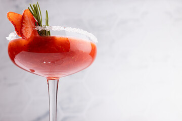 Cold summer strawberry alcohol cocktail margarita with frozen ice, green rosemary twig, berry slices, sugar rim in soft light white modern minimal interior in sunny day, closeup.