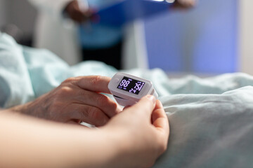 Close up of oxymeter attached on sick senior patient laying in hospital bed, monitoring hemoglobin....