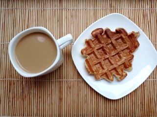 Breakfast. Coffee with milk in a white mug and a waffle on a saucer on a bamboo napkin.