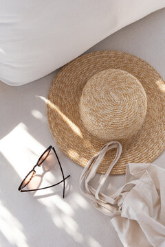 Flatlay of women's fashion accessories. Stylish female sunglasses, straw hat, shopper bag on white lounge couch with pillows. Top view
