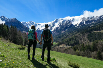 Fototapeta na wymiar A couple with big hiking backpacks holding hands and enjoying the panoramic view on Baeren Valley in Austrian Alps. The highest peaks are snow-capped. Lush green pasture. Sunny day. High mountains
