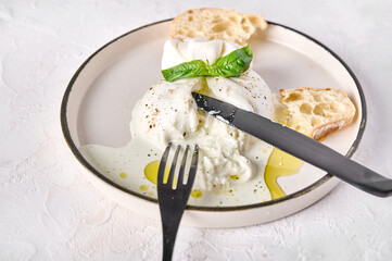 Close up burrata traditional cheese with ciabatta bread and olive oil on white plate with black knife and fork. High key