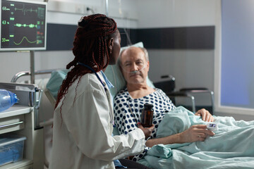 Senior man breathing with help from oxygen tube laying in hospital bed, listening african american doctor explaying diagnosis treatmant holding pills bottle for recovery.