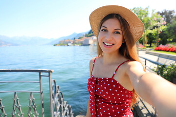 Adorable young woman with hat making selfie photo on Lake Como. Self portrait girl posing in...