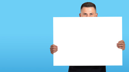 Fototapeta premium Man with banner. Happy man. Person with poster on a blue background. Guy demonstrates positive emotions. Man looks at the camera and covers his face with a poster. A banner with a place for the text.