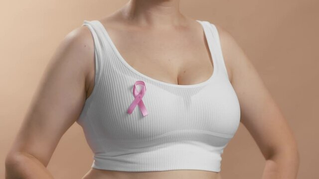 Busty girl in a white top putting a ribbon sign on her chest to support pink October and females fighting against breast cancer. Anonymous slow motion studio shot video on beige background.