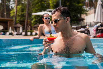 Man holding glass of cocktail in swimming pool near blurred girlfriend at background