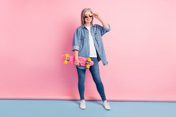 Full size photo of cool aged lady touch eyewear hold skate wear jeans shirt isolated on pink background