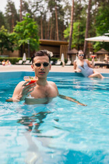 Man in sunglasses holding cocktail in swimming pool near blurred girlfriend