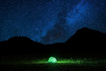 Tent glowing under a starry summer night in the countryside. Tent illuminated by a light. Camping in the mountains under the starry sky. Camping and wild life concept. Real outdoor adventure.