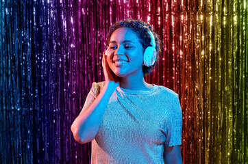 nightlife, technology and people concept - happy young african american woman in headphones listening to music at party in neon lights over rainbow foil fringe curtain background