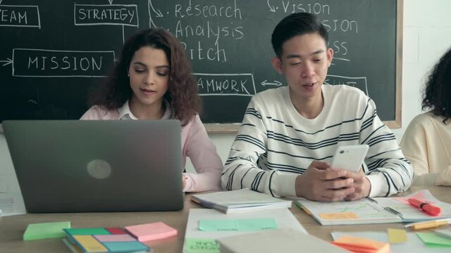 Serious diverse multiracial young college university students business startup group team working on project together learning using devices in classroom office using digital gadgets sitting at desk.