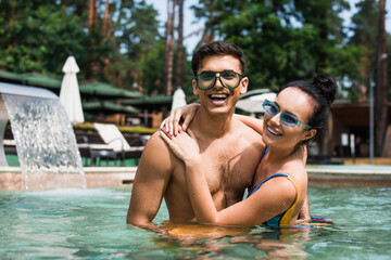 Cheerful couple hugging and looking at camera in swimming pool