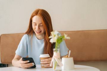 Front view of smiling young woman using smartphone chatting with friends and drinking hot coffee sitting at desk in cozy cafe. Pretty redhead Caucasian lady having leisure activity in coffee shop.
