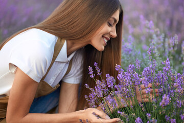 Side view of stunning young smiling woman wearing farm outfit smelling lavender meadow in summer....