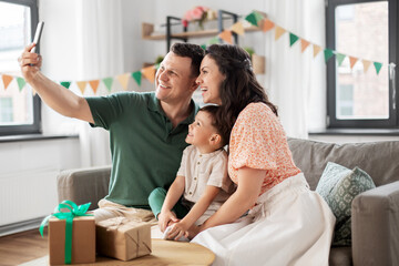 family, holidays and people concept - portrait of happy mother, father and little son with gifts and smartphone taking selfie on birthday at home party