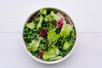Green mix salad in a bowl on white wooden background, top view, flat lay. Healthy food, summer recipes.