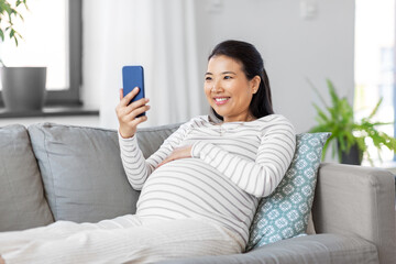 pregnancy, rest, people and expectation concept - happy smiling pregnant asian woman with smartphone sitting on sofa at home