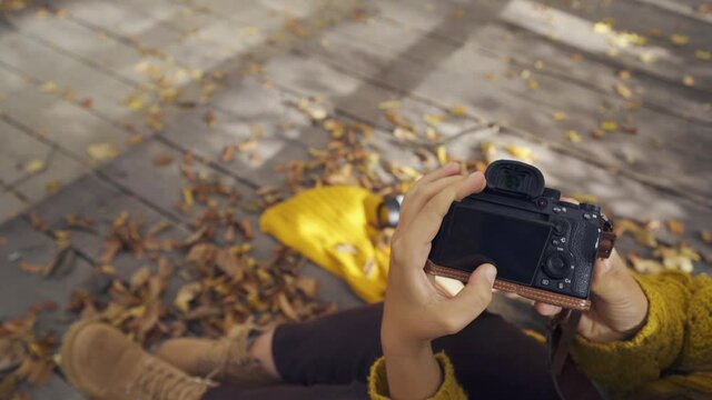 Lady takes picture of open paper book and thermocup on yellow scarf with digital camera on wooden deck bridge with dry leaves in park close view
