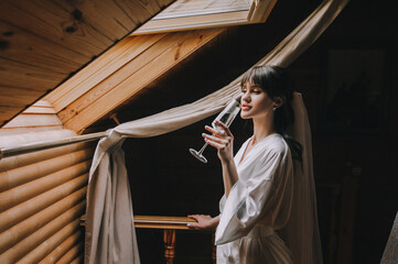 Obraz na płótnie Canvas A beautiful, sweet bride in white lingerie with a long veil stands, posing with a glass of champagne in the wooden room of the hotel near the light window. Wedding photography.