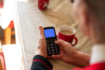 Older women in red sweaters learn to use a large mobile phone. Grandma is sitting at the table and typing on the phone with the keys. There is coffee on the table with the musk